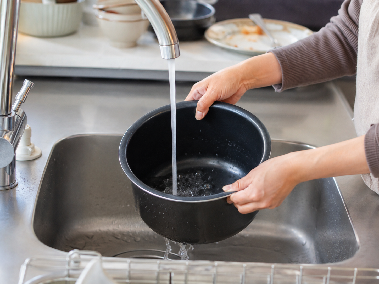 A person rinses a removable black rice-cooker inner pot under the kitchen faucet.