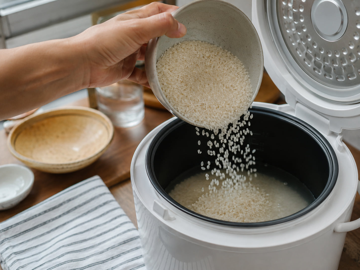 A hand pours uncooked rice into the inner pot of an open, unbranded rice cooker.